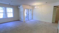 This single-floor residence offers a clean, functional layout with a welcoming and airy atmosphere throughout. The main living area is a generous, open space finished with wall-to-wall plush carpeting that adds warmth and comfort underfoot. Soft, neutral wall tones create a bright and versatile backdrop, complemented by abundant natural light that filters in through multiple windows.

A standout feature of the living area is the elegant chandelier overhead, lending a refined touch to the otherwise understated space. Sliding glass doors with grid-patterned detailing open directly to an outdoor balcony, seamlessly connecting the interior to the exterior and allowing for excellent ventilation and natural light. The balcony offers views of the surrounding area, providing a pleasant outdoor retreat.

The ceiling is well-proportioned, with visible HVAC vents integrated cleanly into the structure, reflecting a practical and well-maintained interior. Recessed structural details near the sliding doors add subtle architectural interest to the layout.

Overall, the space presents a bright, move-in-ready environment with a simple yet polished aesthetic, well-suited for everyday living. The combination of natural light, carpeted floors, and outdoor access makes this a comfortable and inviting single-floor residence.