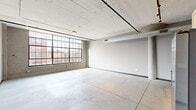 This single-floor loft unit showcases a compelling industrial aesthetic throughout, blending raw architectural elements with modern finishes. Polished concrete floors run continuously across the entire space, complementing the exposed ductwork and painted wood-plank ceilings that lend the unit a distinct warehouse character. Track lighting runs along ceiling rails, providing flexible illumination across the open areas.

The expansive living and dining area is anchored by oversized, steel-framed factory windows that flood the space with natural light and offer views of the surrounding urban landscape. The kitchen features white cabinetry, open shelving, stainless steel appliances including a refrigerator, range, and microwave, along with a wall-mounted HVAC unit overhead.

A long hallway connects the private areas of the unit, where dark-painted walls create a bold contrast to the lighter communal spaces. The hallway also houses a stacked in-unit washer and dryer. The bedroom offers a comfortable footprint with neutral gray walls. Three bathrooms are distributed throughout the unit; one features a subway-tiled tub-shower combination, while another includes a standalone shower, both with marble-topped vanities and concrete tile flooring.