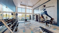 Located at 13645 E Colonial Dr in Orlando, Florida, this single-floor fitness center is designed to inspire and motivate. The space is defined by soaring double-height ceilings and expansive floor-to-ceiling windows that flood the interior with abundant natural light and offer views of the surrounding lush, palm-lined courtyard and neighboring buildings.

The layout is open and well-organized, with distinct zones dedicated to various workout styles. The strength training area features a full dumbbell rack, adjustable weight benches, a cable machine, and a multi-functional power rack, complemented by a punching bag for functional training. A bold, large-scale wall mural declares "The Sport of Fitness Has Arrived," serving as a dynamic visual centerpiece that energizes the entire space.

The cardio zone is equally well-equipped, offering treadmills, spin bikes, a water rowing machine, and a tire-flip sled, catering to a wide range of training preferences. Overhead ceiling fans ensure comfortable airflow throughout.

The entryway features striking teal-blue doors and trim accents that carry through the space, adding a cohesive, branded aesthetic. Durable patterned carpet tiles in neutral tones with blue accents cover the floors throughout. Recessed lighting and pendant fixtures provide even illumination, making this a bright, professional, and fully equipped fitness facility.
