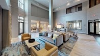 This contemporary amenity lobby is a thoughtfully designed communal space that combines sophisticated architecture with inviting, functional interiors. The entrance features floor-to-ceiling glass double doors flanked by large interior windows, flooding the space with natural light and offering views into the adjoining mail and package area. Soaring double-height ceilings define the expansive main lounge, which is anchored by a sleek, dark-tiled double-sided fireplace — a striking focal point that separates and connects seating zones. The lounge is furnished with a curated mix of neutral sofas, tan leather accent chairs, and layered area rugs in teal and earth tones, all set atop warm wood-look flooring. A standout architectural feature is a textured mosaic wood-clad column near the entry, adding an organic, artisanal contrast to the otherwise modern palette. Adjacent to the lounge, a fully equipped catering kitchen features quartz countertops, herringbone white tile and iridescent mosaic backsplash accents, stainless steel appliances, and pendant lighting. A separate dining nook offers built-in banquette seating, a live-edge wood table, statement pendant lights, and large windows, making it ideal for casual gatherings. Throughout, curated artwork and warm lighting contribute to a refined yet welcoming ambiance.