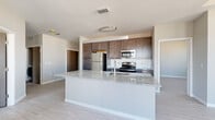 This single-floor unit offers a clean, contemporary design characterized by neutral grey and white tones, light hardwood-style flooring, and crisp white baseboards throughout. The open-concept layout connects the kitchen and living areas seamlessly, creating a spacious, airy feel enhanced by natural light streaming in from a sliding glass door that leads to a private balcony.

The kitchen serves as a central feature, anchored by a large island with a granite countertop and an integrated sink, illuminated by a modern pendant light fixture overhead. Rich dark-toned cabinetry provides generous storage, while stainless steel appliances — including a refrigerator, electric range, and over-the-range microwave — complete the functional setup. A white subway tile backsplash adds a classic touch against the darker cabinetry.

The living area is open and versatile, with ample wall space and good connectivity to adjacent rooms. Bedrooms include wire-rack closet systems offering organized storage, and at least one bedroom features direct access to a bathroom and a dedicated closet area. The bathroom includes dark cabinetry with a vanity sink. Throughout the unit, the consistent use of light flooring, soft wall colors, and modern door finishes creates a cohesive and functional living environment.