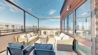 Located at 2005 S 300 W in Salt Lake City, Utah, this rooftop terrace offers an elevated outdoor living experience with panoramic views of the surrounding cityscape and the Wasatch Mountain range. The space is defined by its warm, rich wood-clad exterior walls that add a natural, contemporary aesthetic and serve as a striking architectural backdrop throughout the terrace.

The rooftop is organized into two distinct functional zones. The lounge area features a plush wicker sectional sofa with neutral cushions arranged around a glass-top fire pit table, creating an inviting setting for relaxed gathering. Adjacent lounge chairs add additional seating flexibility. Overhead, café-style string lights are strung across the open-air pergola framework, providing a warm ambiance during evening use.

The dining and grilling zone includes a full-sized stainless steel grill and a large outdoor dining table with seating for six, ideal for al fresco meals. Large windows along the wood-clad wall allow natural light into the interior while maintaining visual connectivity between indoor and outdoor spaces. A glass-panel entry door provides seamless access between the two areas.

The terrace is finished with large-format tile pavers throughout, lending a clean and cohesive look underfoot. Open railings on the perimeter allow unobstructed views of the Salt Lake Valley, making this rooftop an exceptional space for entertaining and relaxation.