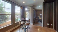 Located in Johns Creek, Georgia, this thoughtfully designed single-floor community space blends modern sophistication with warm, inviting character. The kitchenette area is appointed with rich dark wood cabinetry, striking granite countertops, and an eye-catching white textured tile backsplash. Industrial-style pendant lights hang above the counter, casting a warm glow over the prep and serving areas, which include a sink and coffee station — ideal for hosting gatherings or casual refreshment.

The lounge area features multiple distinct seating zones, each with its own personality. A cozy lounge room showcases a dramatic exposed brick accent wall, plush carpeting, and large windows that frame views of the surrounding community. A wall-mounted large-screen TV anchors the space, complemented by stylish furnishings in neutral and accent tones. An intimate booth-style nook with upholstered bench seating and decorative throw pillows provides a more private retreat.

A casual dining area near corner windows offers abundant natural light and scenic outdoor views, furnished with a high-top bistro table and rustic wood stools. The polished hardwood flooring throughout the common areas adds warmth and continuity. The overall ambiance is urban-chic yet comfortable, making this a well-equipped and welcoming gathering space.