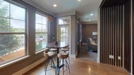 Located in Johns Creek, Georgia, this thoughtfully designed single-floor community space blends modern sophistication with warm, inviting character. The kitchenette area is appointed with rich dark wood cabinetry, striking granite countertops, and an eye-catching white textured tile backsplash. Industrial-style pendant lights hang above the counter, casting a warm glow over the prep and serving areas, which include a sink and coffee station — ideal for hosting gatherings or casual refreshment.

The lounge area features multiple distinct seating zones, each with its own personality. A cozy lounge room showcases a dramatic exposed brick accent wall, plush carpeting, and large windows that frame views of the surrounding community. A wall-mounted large-screen TV anchors the space, complemented by stylish furnishings in neutral and accent tones. An intimate booth-style nook with upholstered bench seating and decorative throw pillows provides a more private retreat.

A casual dining area near corner windows offers abundant natural light and scenic outdoor views, furnished with a high-top bistro table and rustic wood stools. The polished hardwood flooring throughout the common areas adds warmth and continuity. The overall ambiance is urban-chic yet comfortable, making this a well-equipped and welcoming gathering space.