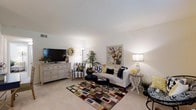 This single-floor residence centers around a thoughtfully designed living room that balances comfort and style. The space features neutral, light-toned walls and smooth ceilings that contribute to an open, airy atmosphere. Plush wall-to-wall carpeting in a warm beige tone runs throughout, softening the overall feel of the room.

The living area is anchored by a crisp white sofa accented with navy and patterned throw pillows, complemented by a matching striped armchair that adds a classic touch. A bold, multicolored geometric area rug defines the seating area and introduces vibrant pops of color. A round black coffee table with gold-toned stools serves as a functional and decorative centerpiece.

A large dresser in white finish doubles as a media console, housing a flat-screen television alongside decorative greenery and accent pieces. Soft ambient lighting is provided by a floor lamp and a decorative table lamp, both contributing to the warm, inviting glow of the room. A colorful floral artwork on the wall adds a lively focal point, while potted plants bring a refreshing natural element to the space.