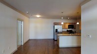 This single-floor space features an open-concept layout that seamlessly connects the living area and kitchen, creating a spacious and functional environment. The kitchen is well-appointed with warm wood cabinetry, granite countertops, and a suite of stainless steel appliances, including a French door refrigerator with an ice and water dispenser and a black range. A central island with a built-in sink provides additional prep space and serves as a natural dividing point between the kitchen and the open living area. Pendant lighting hangs above the island, complemented by track lighting overhead, ensuring the kitchen is well-lit for both cooking and entertaining. The living area is generously sized, offering a blank canvas with neutral-toned walls and rich, dark hardwood flooring that extends throughout the entire space. Recessed and flush-mount ceiling fixtures provide ambient lighting across the open floor plan. A doorway is visible at one end, offering access to additional areas of the unit. The overall ambiance is clean, modern, and move-in ready, with a cohesive design aesthetic anchored by warm wood tones and contemporary finishes.