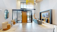 This single-floor space offers a sophisticated, multi-functional layout that blends welcoming social areas with professional meeting facilities. The entry foyer immediately impresses with its dramatic double-height ceiling and expansive floor-to-ceiling black-framed glass walls, flooding the space with natural light and offering open outdoor views. A warm timber pivot door anchors the entrance, complemented by large-format light stone tile flooring that flows throughout.

The main lounge area is anchored by a built-in media wall featuring vertical wood slat detailing and a dark surround, framing a wall-mounted display. Plush sectional sofas in neutral tones with warm accent pillows provide comfortable seating beneath the soaring ceiling. Striking pendant light fixtures, including a cascading gold chainmail chandelier, add an artistic focal point overhead.

A formal conference room accommodates a generous oval wood table surrounded by upholstered chairs, with a textured accent wall displaying a branded gold logo and a grid of circular recessed ceiling lights creating a polished atmosphere. Adjacent flexible seating nooks with glass-paneled sliding partitions offer semi-private areas for smaller gatherings. Fluted wall panels, curated artwork, and cohesive warm-toned furnishings contribute to an overall ambiance that is modern, refined, and inviting.