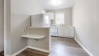 This single-floor unit offers a practical and efficiently designed layout, encompassing a main living area, a kitchen, and a bathroom. The living space features durable wood-look plank flooring in a warm medium-brown tone that runs throughout, complemented by neutral grey walls and white baseboards that lend a clean, modern feel. A ceiling fan with a built-in light fixture is centrally mounted, providing both air circulation and ambient lighting. Windows fitted with blinds allow natural light to filter into the space, and a wall-mounted HVAC unit ensures climate control. The entry door features a windowed panel, adding additional light to the room.

The kitchen is separated from the main living area by a half-wall partition with a fold-down counter surface, creating a functional breakfast bar or workspace. White cabinetry lines the kitchen walls, accompanied by a gas range, refrigerator, and a single-basin sink beneath a window. Light-toned countertops complete the kitchen's clean, cohesive aesthetic, and the same wood-look flooring carries through seamlessly.

The bathroom is compact and straightforward, featuring a walk-in shower stall with a single chrome fixture, a pedestal sink, and a mirrored medicine cabinet, all finished in bright white for a crisp appearance.