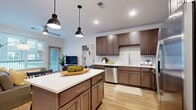 Located at 1879 Village West Pkwy in Kansas City, KS, this thoughtfully designed one-bedroom, one-bathroom residence offers a comfortable and modern living experience across a single floor. Light-toned hardwood-style flooring runs throughout the main living areas, creating a cohesive and airy aesthetic.

The open-concept kitchen is a standout feature, boasting rich dark-brown shaker cabinetry, white quartz countertops, a subway tile backsplash, and a full suite of stainless steel appliances including a refrigerator with ice maker, range, microwave, and dishwasher. A central island with pendant lighting provides additional prep space and casual seating.

The dining area is bathed in natural light from a large window with outdoor views, offering a welcoming space for meals. The living room features a sliding glass door that opens to the exterior, allowing abundant natural light to fill the room.

The bedroom is a serene retreat with plush carpet flooring, a ceiling fan, and access to a dedicated closet. The bathroom is well-appointed with a dark-toned vanity, white quartz countertop, wood-look tile flooring, and a glass-enclosed walk-in shower with white tile surround.