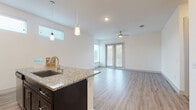 This single-floor unit offers a clean, contemporary design with a bright, open-concept living and kitchen area at its core. The kitchen is well-appointed with dark espresso cabinetry, granite countertops, a full suite of stainless steel appliances including a refrigerator, microwave, and range, and a peninsula island with an undermount sink and elegant pendant lighting overhead. Elevated windows along the kitchen wall allow natural light to filter in without compromising wall space. The adjoining living area is generously sized, finished with light wood-style plank flooring, recessed lighting, and a ceiling fan. French doors at the far end of the living space provide access to the outdoors and invite additional natural light into the room. A hallway leads from the main living area to the private quarters, including a bedroom finished with plush carpet flooring, a ceiling fan, and a window with blinds. The bathroom features a granite-topped vanity with dark cabinetry, a glass-enclosed walk-in shower with tiled surround, and tile flooring. A walk-in closet adjoins the bathroom, offering practical storage. Throughout, the unit maintains a consistent, neutral color palette that enhances its modern, move-in-ready appeal.