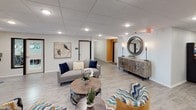 This single-floor lobby and common area serves as the welcoming heart of Regency Multifamily, offering residents and guests a polished, thoughtfully designed space from the moment they arrive. The entrance features a glass vestibule with black-framed doors, a branded welcome mat, and views into an adjacent office or leasing area, establishing a professional first impression.

Beyond the entry, the interior opens into a generously proportioned lounge that combines comfort with contemporary style. Light wood-look flooring flows throughout, complemented by a drop tile ceiling with recessed lighting that provides consistent, even illumination. Accent walls in warm amber tones add energy and visual interest to the neutral color palette.

The lounge is furnished with a curated mix of seating — including a gray sofa with navy accent pillows, patterned accent chairs in teal and gold chevron, and a round wood coffee table — all arranged to encourage relaxed social interaction. Decorative touches such as a large ornate round mirror, a rustic wood-and-metal sideboard with floor lamps, and nature-inspired artwork lend the space a refined yet approachable character.

A dedicated coffee and refreshment station with a clock above it adds everyday convenience for residents. Directional signage guides visitors toward unit corridors, making the space both functional and welcoming.