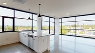 Located at 309 E 15th St in Vancouver, Washington, this contemporary single-floor residence showcases a refined modern design with clean lines and a bright, open aesthetic throughout. The standout feature of the living and kitchen area is the expansive floor-to-ceiling window system framed in bold black, wrapping around the corners of the space to deliver sweeping panoramic views of the Vancouver skyline and surrounding treetops, flooding the interior with abundant natural light.

The open-concept kitchen is well-appointed with a large central island featuring a built-in sink and pendant lighting, white cabinetry, stainless steel appliances including a gas range, microwave, and side-by-side refrigerator, and a hexagonal tile backsplash that adds subtle texture. Polished concrete flooring runs throughout the main living areas, contributing to the unit's contemporary industrial character.

A clean, elongated hallway connects the private areas of the residence, where carpeted bedrooms offer a softer contrast to the polished common spaces. Two bathrooms are finished with floating white vanities, stone-look countertops, backlit mirrors, and black hardware accents, maintaining a consistent modern palette. The overall ambiance is minimal, bright, and polished, making excellent use of natural light and elevated urban views.