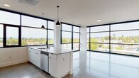 Located at 309 E 15th St in Vancouver, Washington, this contemporary single-floor residence showcases a refined modern design with clean lines and a bright, open aesthetic throughout. The standout feature of the living and kitchen area is the expansive floor-to-ceiling window system framed in bold black, wrapping around the corners of the space to deliver sweeping panoramic views of the Vancouver skyline and surrounding treetops, flooding the interior with abundant natural light.

The open-concept kitchen is well-appointed with a large central island featuring a built-in sink and pendant lighting, white cabinetry, stainless steel appliances including a gas range, microwave, and side-by-side refrigerator, and a hexagonal tile backsplash that adds subtle texture. Polished concrete flooring runs throughout the main living areas, contributing to the unit's contemporary industrial character.

A clean, elongated hallway connects the private areas of the residence, where carpeted bedrooms offer a softer contrast to the polished common spaces. Two bathrooms are finished with floating white vanities, stone-look countertops, backlit mirrors, and black hardware accents, maintaining a consistent modern palette. The overall ambiance is minimal, bright, and polished, making excellent use of natural light and elevated urban views.