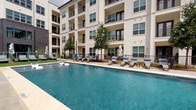 This outdoor pool area serves as a central amenity within a multi-story residential community, offering a thoughtfully designed space for relaxation and recreation. The expansive swimming pool features crystal-clear blue water and is complemented by a separate spa or hot tub area, providing options for both active and leisurely use.

The pool deck is finished with smooth concrete surrounds and is lined with an array of chaise lounge chairs and patio furniture, ideal for sunbathing or unwinding outdoors. Market umbrellas are positioned throughout the deck, offering shaded seating options. Decorative landscaping, including ornamental grasses, planters, and young trees, adds a natural, refreshing element to the environment.

A decorative patterned accent wall enhances the visual appeal of one perimeter section, while iron fencing provides both security and an open sightline to the surrounding greenery. The pool area is accessed through a dedicated entrance with wooden doors, accompanied by posted pool rules and a covered transition area.

The space is surrounded on multiple sides by the residential building's facade — a mix of modern white siding, brick, and large balconied windows — creating a semi-enclosed courtyard atmosphere that feels private while remaining open to natural light and open skies above.