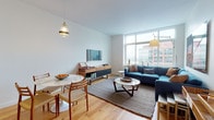 This thoughtfully designed single-floor residence offers a seamless blend of contemporary style and urban living. The open-plan living and dining area is anchored by floor-to-ceiling windows that flood the space with natural light and frame striking city skyline views. Light hardwood flooring flows throughout, complementing the clean white walls and modern finishes. The living area comfortably accommodates a large sectional sofa, a round marble coffee table, and a media console, creating an inviting yet functional layout. A dedicated dining space sits adjacent, featuring a round marble-top table and mid-century modern chairs.

The kitchen is outfitted with a two-tone cabinetry scheme — combining rich wood-grain lower cabinets with crisp white upper cabinets — alongside light tile flooring, a kitchen island with a white countertop, and a full suite of stainless steel appliances including a gas range, microwave, dishwasher, and refrigerator.

Both bedrooms are bright and well-proportioned, each featuring hardwood floors and large windows with urban views. The primary bedroom offers ample space for a full-size bed and dresser. The bathroom is finished with neutral large-format floor tiles, a soaking tub with shower, and clean white walls accented by framed artwork.