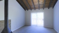 This single-story space showcases a striking architectural centerpiece: a vaulted ceiling with exposed dark wood beams set against warm natural wood planking, lending the interior a rustic yet refined character. The angled ceiling adds a sense of height and openness, making the main living area feel generously spacious.

The room is finished with soft, neutral-toned wall-to-wall carpeting that complements the warm tones of the ceiling above. Clean white walls provide a bright, versatile backdrop throughout the space.

A full-width sliding glass door serves as the room's focal point, drawing in an abundance of natural light and offering a direct view of an outdoor area, likely a balcony or patio, with greenery visible beyond. Vertical blinds offer privacy and light control as needed.

The overall ambiance is bright, airy, and inviting, with the combination of natural wood overhead and soft flooring underfoot creating a comfortable, casual atmosphere. The open, uncluttered layout lends itself well to flexible furniture arrangements, making the space highly adaptable to various living needs.