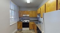 This single-floor residential unit offers a practical and comfortable layout across several well-defined living spaces. The kitchen is efficiently designed in an L-shaped configuration, featuring warm honey-toned wood cabinetry, dark granite-style countertops, a stainless steel sink, and a full suite of appliances including an electric range with overhead ventilation hood and a refrigerator. Tile flooring and a bright window with blinds complete the kitchen's functional character.

The living and bedroom areas are finished with neutral-toned carpet and painted in soft grey throughout, giving the space a calm, cohesive feel. Exposed structural ceiling beams add a distinctive architectural element, while baseboard heating units line the walls for efficient climate control. Multiple windows fitted with blinds allow natural light to filter into the rooms, creating an airy atmosphere. One bedroom benefits from a notably tall, vaulted ceiling and includes a built-in closet, with direct access to a full bathroom.

The bathroom is cleanly appointed with a single-sink vanity topped with a mirrored medicine cabinet, a tub and shower combination, and tile flooring. A second bathroom of similar design provides additional convenience. Overall, the unit presents a straightforward, move-in-ready layout well-suited for everyday living.