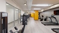 Located at 629 Euclid Ave in Cleveland, Ohio, this fitness facility spans a generously sized single floor divided into three dedicated exercise rooms. The space is designed with a clean, modern aesthetic, featuring light wood-style flooring, white and gray walls accented by bold yellow structural columns that add a striking visual identity throughout.

The main exercise area offers an extensive lineup of strength training machines from True Fitness, including cable systems, chest press, and leg extension equipment, all arranged along the length of the room for an open, navigable layout. Glass partition walls define separate zones while maintaining visual connectivity across the space. Overhead, exposed utility runs and a mix of recessed and flush-mount lighting provide a functional, well-lit environment.

The cardio section is outfitted with treadmills, ellipticals, stationary bikes, and a rowing machine, complemented by wall-mounted flat-screen TVs for entertainment during workouts. A dedicated functional training area houses stability balls, medicine balls, resistance bands, and dumbbells neatly organized on a wall-mounted rack, supporting a variety of training modalities. Storage lockers and sanitation stations are conveniently positioned throughout the facility. The overall ambiance is energetic yet organized, making this a comprehensive fitness destination.