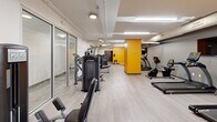 Located at 629 Euclid Ave in Cleveland, Ohio, this fitness facility spans a generously sized single floor divided into three dedicated exercise rooms. The space is designed with a clean, modern aesthetic, featuring light wood-style flooring, white and gray walls accented by bold yellow structural columns that add a striking visual identity throughout.

The main exercise area offers an extensive lineup of strength training machines from True Fitness, including cable systems, chest press, and leg extension equipment, all arranged along the length of the room for an open, navigable layout. Glass partition walls define separate zones while maintaining visual connectivity across the space. Overhead, exposed utility runs and a mix of recessed and flush-mount lighting provide a functional, well-lit environment.

The cardio section is outfitted with treadmills, ellipticals, stationary bikes, and a rowing machine, complemented by wall-mounted flat-screen TVs for entertainment during workouts. A dedicated functional training area houses stability balls, medicine balls, resistance bands, and dumbbells neatly organized on a wall-mounted rack, supporting a variety of training modalities. Storage lockers and sanitation stations are conveniently positioned throughout the facility. The overall ambiance is energetic yet organized, making this a comprehensive fitness destination.