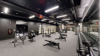 Located at 1950 Broadway in Oakland, CA, this large-scale fitness facility delivers a comprehensive, professionally designed training environment across a single expansive floor. The space is defined by an industrial-modern aesthetic, with exposed black ceilings revealing structural ductwork and mechanical systems, contrasted by bright linear LED lighting that ensures excellent visibility throughout.

The floor plan is organized into several distinct training zones. A dedicated strength area features multiple barbell platforms, squat racks, weight trees, and adjustable benches suited for free-weight training. Adjacent to this, a generous machine circuit area is outfitted with rows of black selectorized strength machines arranged around structural columns, which are accented with rope wrapping for a refined industrial touch.

A standout feature is a long sprint and functional training lane, marked with orange and white lines on dark rubberized flooring. Along one wall, a bold orange MoveStrong functional rig system spans the length of the lane, integrating monkey bars, wall-mounted ladders, medicine ball storage, barbell holders, and resistance band stations. A glass-enclosed studio with hardwood flooring provides a dedicated space for classes or flexibility work. Full-length mirrors throughout enhance the sense of openness. Rubber and carpet-tile flooring unify the space while providing safety and performance underfoot.