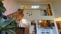 This two-story home showcases an open and airy design anchored by a dramatic vaulted ceiling with a centrally placed skylight that floods the main living area with abundant natural light. A classic wooden staircase with turned spindle railings connects the two floors, while an open upper-level landing overlooks the main living space below, creating a sense of vertical openness and connectivity throughout the home. Warm wood tones are carried through the stair railings and cabinetry, lending a cohesive and inviting character to the interior. Decorative hanging planters and lush indoor greenery add a natural, lived-in charm to the space. The kitchen is positioned adjacent to the main living area and features warm wood cabinetry, a white refrigerator, and a microwave, with ample counter space and storage. A ceiling fan provides comfortable air circulation in the lower living area. The layout promotes a smooth flow between the kitchen, dining, and living spaces, making the home well-suited for both everyday living and entertaining. The combination of skylights, open railings, and multi-level design gives the home a bright, spacious feel that is both functional and visually engaging.