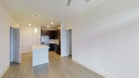 This single-floor residence showcases a clean, modern design with an open-concept layout that seamlessly connects the kitchen and living areas. The kitchen is a standout feature, equipped with stainless steel appliances including a refrigerator, microwave, and range, complemented by sleek dark cabinetry that contrasts beautifully against light countertops. A white kitchen island with an integrated sink serves as a functional centerpiece, ideal for meal preparation or casual dining. Pendant lighting above the island adds a stylish touch, while recessed ceiling lights provide consistent illumination throughout the space. Light-toned hardwood flooring runs throughout the unit, lending warmth and a sense of continuity to the open layout. The living area is bright and airy, featuring neutral white walls and a ceiling fan that enhances comfort. A hallway visible from the main living space leads to additional rooms, offering a well-organized flow. The overall ambiance is contemporary and welcoming, with a thoughtful balance of form and function that makes this space both practical and visually appealing.