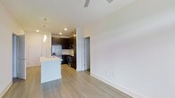 This single-floor residence showcases a clean, modern design with an open-concept layout that seamlessly connects the kitchen and living areas. The kitchen is a standout feature, equipped with stainless steel appliances including a refrigerator, microwave, and range, complemented by sleek dark cabinetry that contrasts beautifully against light countertops. A white kitchen island with an integrated sink serves as a functional centerpiece, ideal for meal preparation or casual dining. Pendant lighting above the island adds a stylish touch, while recessed ceiling lights provide consistent illumination throughout the space. Light-toned hardwood flooring runs throughout the unit, lending warmth and a sense of continuity to the open layout. The living area is bright and airy, featuring neutral white walls and a ceiling fan that enhances comfort. A hallway visible from the main living space leads to additional rooms, offering a well-organized flow. The overall ambiance is contemporary and welcoming, with a thoughtful balance of form and function that makes this space both practical and visually appealing.