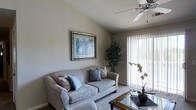 This single-story space showcases a thoughtfully designed living area that balances comfort and functionality. The room features vaulted ceilings that enhance the sense of openness, complemented by a ceiling fan that ensures year-round comfort. Neutral-toned walls in warm greige create a calm and inviting atmosphere throughout.

The living area is anchored by a plush, light grey sofa adorned with decorative accent pillows in soft blue and beige tones, arranged around a glass-topped coffee table with a dark wood frame. A textured area rug defines the seating space and adds warmth to the flooring.

Natural light is a standout feature, flooding the room through large sliding glass doors dressed with vertical blinds, which also provide a glimpse of the outdoors. Lush indoor plants, including a tall decorative tree and a smaller flowering plant near the door, bring a refreshing natural element to the interior. A framed landscape artwork on the wall adds a subtle artistic touch. The textured ceiling adds a classic finish, rounding out this well-appointed and welcoming living space.