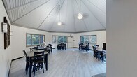 Located at 3215 West Colony Drive in Greenfield, Wisconsin, this single-floor community space offers a well-designed layout ideal for gatherings and events. The centerpiece of the space is a striking octagonal main hall distinguished by a soaring vaulted ceiling with exposed beam detailing and white-painted tongue-and-groove panels. Pendant ceiling fans hang from the apex, and large sliding windows wrap around the perimeter, flooding the room with natural light and offering views of the surrounding mature tree canopy. Light wood-look flooring runs throughout, lending a clean, cohesive aesthetic. The main hall is furnished with multiple dining tables and navy blue chairs, providing ample seating for group events. Adjacent to the main hall is a transitional area featuring warm wood cabinetry with granite-style countertops and recessed lighting — well-suited for serving or buffet-style setups. A fully equipped galley-style kitchenette provides additional functionality, complete with wood cabinetry, granite countertops, a refrigerator, range, and track lighting overhead. Decorative artwork accents the neutral white walls throughout, adding warmth to the space. The layout flows efficiently, with clearly marked exits ensuring safety and accessibility.