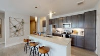This thoughtfully designed single-floor residence offers a cohesive blend of modern finishes and comfortable living spaces. The open-concept kitchen is a standout feature, showcasing rich dark cabinetry, light quartz countertops, a subway tile backsplash, and a full suite of stainless steel appliances. A generously sized kitchen island with an undermount sink and pendant lighting above serves as both a functional workspace and a casual dining area with bar seating. Light wood-tone flooring flows throughout the kitchen and common areas, lending a clean, contemporary feel. The living room is bright and inviting, featuring a ceiling fan, large windows that welcome natural light, and a warm accent wall that adds character to the space. A versatile secondary sitting room with an additional window provides flexible use as a home office or den. The bedrooms are carpeted for comfort, each equipped with ceiling fans, ample natural light, and generous closet space. The primary bedroom connects to a well-appointed en-suite bathroom featuring a dual-sink vanity, tile flooring, and a spacious walk-in closet with built-in shelving and hanging storage. A second full bathroom offers a tub-shower combination with white tile surround. Throughout the residence, neutral tones and clean architectural lines create a calm, polished ambiance.