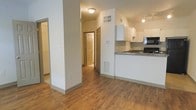 This single-floor residence offers a clean and functional open-concept layout that seamlessly connects the living area and kitchen. The space is finished with warm hardwood-style flooring throughout, providing a cohesive and inviting atmosphere.

The kitchen is well-appointed with white cabinetry, dark stone-finish countertops, and black appliances, including a range and microwave. A peninsula-style counter defines the kitchen space while keeping it visually connected to the main living area. The kitchen is also equipped with a ventilation hood and ample cabinet storage.

A central structural column adds an architectural element to the open living space, subtly defining zones without disrupting the open flow. Flush-mount ceiling lighting provides consistent illumination throughout.

A hallway extends from the main living area, leading to additional rooms and a bathroom, with HVAC vents integrated neatly into the walls and ceiling. The neutral white walls and light-toned trim throughout the space lend a bright, clean aesthetic that is easy to personalize. A smart thermostat is visibly mounted, reflecting modern convenience features built into the home.