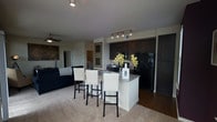 This single-floor residence offers a thoughtfully designed open-concept layout that integrates the kitchen, dining, and living areas into one cohesive and functional space. The kitchen is a standout feature, boasting rich dark cabinetry, black appliances including a full-size refrigerator and range, and a granite-topped island with a breakfast bar accommodating multiple bar stools — perfect for casual dining or entertaining. Track lighting above the kitchen provides focused illumination, complementing the overall modern aesthetic. The transition from hardwood-style flooring in the kitchen to plush carpeting in the living area creates a natural distinction between spaces while maintaining an open, airy feel. The living area is anchored by a ceiling fan and offers a comfortable, welcoming ambiance. Framed artwork along the walls adds a refined touch to the neutral color palette throughout. A hallway visible from the main living space suggests additional rooms beyond, contributing to the overall sense of a well-proportioned and versatile floor plan. The combination of contemporary finishes, practical design, and connected living spaces makes this residence both functional and visually appealing.