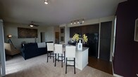 This single-floor residence offers a thoughtfully designed open-concept layout that integrates the kitchen, dining, and living areas into one cohesive and functional space. The kitchen is a standout feature, boasting rich dark cabinetry, black appliances including a full-size refrigerator and range, and a granite-topped island with a breakfast bar accommodating multiple bar stools — perfect for casual dining or entertaining. Track lighting above the kitchen provides focused illumination, complementing the overall modern aesthetic. The transition from hardwood-style flooring in the kitchen to plush carpeting in the living area creates a natural distinction between spaces while maintaining an open, airy feel. The living area is anchored by a ceiling fan and offers a comfortable, welcoming ambiance. Framed artwork along the walls adds a refined touch to the neutral color palette throughout. A hallway visible from the main living space suggests additional rooms beyond, contributing to the overall sense of a well-proportioned and versatile floor plan. The combination of contemporary finishes, practical design, and connected living spaces makes this residence both functional and visually appealing.