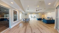 Located at 3924 Calibre Bend Lane in Winter Park, Florida, this thoughtfully designed single-story community clubhouse offers a welcoming and functional environment across multiple interconnected living areas. Wide-plank light hardwood flooring flows seamlessly throughout, unifying the space with a clean, contemporary aesthetic.

The main gathering area is expansive and open, featuring tray ceilings with recessed lighting and a modern ceiling fan, creating an airy, well-lit atmosphere. A dining table with sleek chairs sits near large glass sliding doors that invite abundant natural light and provide views of the outdoor surroundings. A dedicated lounge area features two mid-century modern navy blue sofas arranged around a coffee table, anchored by a striking white textured brick accent wall.

Additional seating nooks with bar-height tables and modern stools offer casual gathering spots throughout. A standout business center showcases vibrant teal brick-paneled workstation alcoves, bathed in natural light from large windows overlooking lush greenery. Community bulletin boards and a "Pet of the Month" display add a personable, neighborly character. A staff office and accessible restroom facilities are also present. The overall ambiance is bright, modern, and inviting, designed to serve as a true community hub.