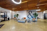 Located at 3118 6th Ave in Tacoma, Washington, this contemporary lobby space makes a strong architectural statement from the moment you enter. The open-plan layout is defined by a warm wood slat ceiling with integrated linear lighting, which creates a rich contrast against the polished concrete floors below. Large black-framed glass entry doors and floor-to-ceiling glazing invite abundant natural light and provide clear sightlines to the street, enhancing the sense of openness.

A prominent exposed concrete accent wall serves as the visual centerpiece, featuring an oversized ampersand graphic that adds a bold, artistic touch to the space. Lush tropical plants are strategically positioned throughout, softening the industrial materials and adding life and color. The seating area is furnished with a patterned modular sofa, sleek metal-framed leather chairs, and minimalist side tables, creating a comfortable and stylish gathering zone.

Additional features include a stainless steel elevator, a wall-mounted display screen near the entry, a decorative wooden art installation, and a water fountain station. The overall ambiance blends industrial and modern design elements — raw concrete, natural wood, and clean white walls — resulting in a sophisticated and inviting communal space well-suited for residents or visitors.