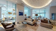This expansive single-floor amenity space is designed with a refined, contemporary aesthetic that seamlessly blends artistic detail with functional elegance. The centerpiece of the main lounge area is a dramatic oval ceiling installation adorned with cascading sculptural butterfly-like elements, backlit to create a warm, ambient glow that anchors the entire room. Beneath it, intricate parquet wood flooring in a geometric star pattern extends throughout, adding warmth and visual depth to the open layout.

Multiple distinct seating zones are thoughtfully arranged throughout the space. Plush sectional sofas, lounge chairs, and accent tables create intimate gathering areas, while floor lamps and curated artwork — including bold abstract canvases — contribute to the sophisticated atmosphere. A cozy nook with built-in upholstered bench seating and a marble-topped round table offers a quieter retreat.

The kitchen and dining area features soaring floor-to-ceiling windows overlooking an outdoor terrace and the surrounding cityscape, flooding the space with abundant natural light. A substantial marble-topped island with integrated gas cooktop anchors the kitchen, accompanied by a long communal dining table with modern upholstered chairs. Additional seating areas with swivel chairs and a games area, including a billiard table, round out this versatile, amenity-rich environment.