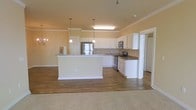 Located at 1850 Popps Ferry Rd in Biloxi, Mississippi, this single-floor residence offers a well-designed, open-concept layout that seamlessly connects the kitchen and living areas. The kitchen is a standout feature, outfitted with white cabinetry, dark granite-style countertops, and stainless steel appliances including a refrigerator, range, and microwave. A large central island provides generous prep space and serves as a natural gathering point. Pendant lighting above the island and a chandelier near the adjacent living area add warm, decorative illumination throughout the open space. Crown molding along the ceilings lends an elegant, finished touch to the interior. The flooring transitions from rich hardwood-style planks in the kitchen and common areas to plush carpet in the living space, offering both practicality and comfort. The warm, neutral color palette on the walls creates a bright and welcoming ambiance throughout. The open floor plan maximizes the sense of space, making the home feel airy and functional. This single-story layout provides convenient, accessible living with cohesive design elements that make it both practical and inviting.