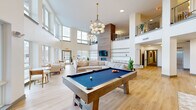 Located at 195 Mansfield Ave in Norton, Massachusetts, this generously sized two-floor amenity space offers a well-appointed environment designed for social gathering and community engagement. The ground floor serves as the primary hub, featuring soaring double-height ceilings flooded with natural light from expansive floor-to-ceiling windows. Light hardwood flooring runs throughout, complementing the bright white walls and creating a warm, modern aesthetic. A statement fireplace with a wood-paneled surround anchors the main lounge area, where plush neutral seating arrangements invite relaxation. A billiards table and shuffleboard table provide recreational focal points, while globe and sputnik-style chandeliers add a stylish finishing touch. A built-in bar area with blue tile backsplash and a mailbox alcove reflect the space's community-oriented function. An elegant staircase with metal railings connects to the upper floor, which offers additional open lounge and seating areas bathed in natural light. The upper level features its own distinct chandelier accents, open sightlines to the floor below via a mezzanine-style overlook, and corridors leading to further amenity spaces. Together, both floors create a cohesive, thoughtfully designed environment suited for community living.