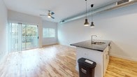 Located in the vibrant Carytown neighborhood of Richmond, Virginia, this single-floor unit blends modern industrial design with contemporary finishes. Rich, variegated hardwood flooring runs throughout the space, lending warmth and character to every room. The open-concept living and kitchen area serves as the heart of the unit, where an expansive granite-topped island with an integrated sink anchors the layout. The kitchen is outfitted with stainless steel appliances, white subway tile backsplash, and crisp white cabinetry, all illuminated by stylish pendant lighting. An exposed metallic ductwork element runs across the ceiling, adding an industrial-chic aesthetic that complements the clean, neutral walls. A ceiling fan and sliding glass door in the living area invite natural light and provide access to a private outdoor balcony with views of surrounding greenery. The bathroom features a full soaking tub with a tiled surround, accented by an elegant mosaic tile inlay and brushed nickel fixtures. A dedicated laundry area with a stacked washer and dryer adds everyday convenience. The overall ambiance is bright, open, and thoughtfully designed for comfortable urban living.