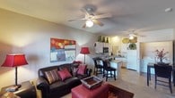 This single-floor space showcases an open-concept layout where the living area and kitchen connect naturally, creating a cohesive and functional environment. The living room is anchored by a dark leather sofa adorned with colorful accent pillows, a red upholstered ottoman serving as a coffee table, and a floor lamp providing warm ambient lighting. A vibrant piece of wall art adds a bold pop of color, complementing the overall décor. Ceiling fans in both the living and kitchen areas ensure comfortable airflow throughout.

The kitchen is designed for practicality, featuring white cabinetry, a built-in microwave, and a white refrigerator. A peninsula-style breakfast bar with dark bar stools creates a casual dining option while maintaining the open flow of the space. Decorative vases and woven wall accents add personality to the kitchen area. Tile flooring in the kitchen transitions to carpeting in the living room, subtly defining each zone. The textured ceiling and neutral wall tones provide a warm, understated backdrop throughout the space.