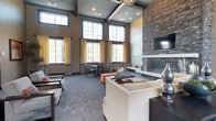 This elegant single-floor community lounge makes an immediate impression through its grand entrance lobby, where large-format tile flooring, stacked stone columns, and pendant lighting set a refined tone. Floor-to-ceiling glass doors flood the entry with natural light, creating a welcoming first impression.

The main lounge area is defined by dramatically high ceilings with exposed dark wood beams and an expansive arrangement of tall, multi-pane windows dressed in layered gold and gray curtains. A floor-to-ceiling stacked stone accent wall serves as the room's focal point, anchoring a sleek linear fireplace topped by a wall-mounted television. Comfortable seating—including neutral sofas, accent armchairs, and vibrant orange ottomans—encourages relaxed gathering throughout the space.

A dedicated dining area features a substantial wooden table with upholstered, nail-head-trimmed chairs, ideally positioned beneath the oversized windows to take full advantage of the natural light and exterior views. Additional casual seating with a round table provides a more intimate setting nearby.

A built-in dark wood media and shelving unit adds functionality and visual interest to one corner of the lounge. The kitchenette area is outfitted with dark cabinetry, a mosaic tile backsplash, stainless steel appliances, a compact refrigerator, and a coffee station, making the space well-suited for events and everyday use alike.