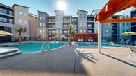 Located at 3751 Heritage Towne Blvd in Midlothian, TX, this generously sized outdoor pool area offers a resort-style amenity experience within a modern residential community. The space encompasses a large, freeform swimming pool with crystal-clear blue water, complemented by integrated water features including fountain jets that add both visual appeal and a relaxing ambiance. A separate wading area and spa provide additional options for leisure and relaxation.

The pool deck is finished in a textured aggregate concrete surface, providing ample space for lounging and socializing. Multiple chaise lounges and chairs are arranged throughout the deck, shaded in part by vibrant yellow umbrellas. A standout feature is the substantial wood-framed pavilion, or cabana, which provides a shaded gathering area with seating underneath its pitched roof — ideal for group activities or escaping the Texas sun.

Decorative tall palm trees are strategically placed throughout the pool area, reinforcing the resort-like atmosphere. The entire perimeter is enclosed by a black iron security fence, ensuring safety and privacy. The backdrop of the multi-story residential buildings, constructed with a contemporary mix of brick, stucco, and dark accent panels, frames the space and contributes to its vibrant, community-centered character. The open-sky setting maximizes natural light throughout the day.