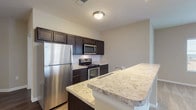 Located at 40 Hutchinson Ave in Columbus, Ohio, this single-floor unit offers a thoughtfully designed layout with modern finishes throughout. The open-concept kitchen is a standout feature, showcasing rich dark cabinetry, light granite-style countertops, a classic subway tile backsplash, and a full suite of stainless steel appliances including a refrigerator, range, and over-the-range microwave. A peninsula with a matching countertop provides additional workspace and serves as a natural divider between the kitchen and the main living area. The living space is generously sized, finished with plush neutral carpeting, and bathed in natural light from large windows and a glass door that opens to a private balcony. Track lighting adds a contemporary touch to the living area. The unit includes two well-sized bathrooms, each featuring dark vanity cabinetry, granite-style countertops, and durable wood-look vinyl flooring. The primary bathroom offers a walk-in shower with sliding glass doors. Storage is a strong suit of this unit, with multiple walk-in closets outfitted with wire shelving and hanging systems, providing ample organizational space. The overall aesthetic is clean and contemporary, with neutral warm tones creating a welcoming ambiance throughout.