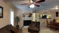 This single-floor residence offers a warm and inviting living space designed for both comfort and functionality. The living room serves as the heart of the home, anchored by a classic white-surround fireplace with a brick hearth, adding a timeless focal point to the space. Crown molding along the ceiling adds a refined architectural touch, while a five-blade ceiling fan with a light fixture provides both practical airflow and ambient lighting. The neutral-toned walls and light carpeting create a calm, cohesive atmosphere throughout. Natural light filters in through a window on the side wall, brightening the room and enhancing its open feel. The living area flows seamlessly into the kitchen, which is visible through a pass-through opening. The kitchen features yellow cabinetry and dark countertops, giving it a distinctive, warm character. The open-concept layout between the living and kitchen areas promotes easy interaction and a connected feel. Decorative elements, including wall art and greenery, contribute to the overall cozy and well-appointed ambiance of the home, making it a welcoming single-level living environment.
