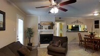 This single-floor residence offers a warm and inviting living space designed for both comfort and functionality. The living room serves as the heart of the home, anchored by a classic white-surround fireplace with a brick hearth, adding a timeless focal point to the space. Crown molding along the ceiling adds a refined architectural touch, while a five-blade ceiling fan with a light fixture provides both practical airflow and ambient lighting. The neutral-toned walls and light carpeting create a calm, cohesive atmosphere throughout. Natural light filters in through a window on the side wall, brightening the room and enhancing its open feel. The living area flows seamlessly into the kitchen, which is visible through a pass-through opening. The kitchen features yellow cabinetry and dark countertops, giving it a distinctive, warm character. The open-concept layout between the living and kitchen areas promotes easy interaction and a connected feel. Decorative elements, including wall art and greenery, contribute to the overall cozy and well-appointed ambiance of the home, making it a welcoming single-level living environment.