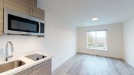 This single-floor studio unit offers a clean, contemporary design throughout, characterized by light-toned luxury vinyl plank flooring, crisp white walls, and recessed lighting that collectively create a bright and functional living environment.

The open-plan living and kitchen area anchors the unit. The kitchenette is fitted with warm wood-toned cabinetry, white countertops, a built-in electric cooktop, a stainless steel over-the-range microwave, and a compact sink — providing all essential cooking amenities in an efficient footprint. A large window in the adjoining living area fills the space with natural light and offers views of the surrounding outdoor scenery.

The sleeping area is a well-proportioned, light-filled room with the same consistent flooring and neutral color palette. A wall-mounted thermostat and control panel are conveniently positioned, contributing to the room's functional design.

The bathroom is neatly appointed with a wall-mounted pedestal sink, a framed mirror with built-in storage, a standard toilet, and a towel bar — all finished in clean white tones with chrome fixtures. The continuous vinyl plank flooring extends into this space, maintaining design consistency throughout the unit.

Overall, this studio presents a modern, move-in-ready space that prioritizes clean aesthetics, practicality, and efficient use of space.