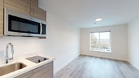 This single-floor studio unit offers a clean, contemporary design throughout, characterized by light-toned luxury vinyl plank flooring, crisp white walls, and recessed lighting that collectively create a bright and functional living environment.

The open-plan living and kitchen area anchors the unit. The kitchenette is fitted with warm wood-toned cabinetry, white countertops, a built-in electric cooktop, a stainless steel over-the-range microwave, and a compact sink — providing all essential cooking amenities in an efficient footprint. A large window in the adjoining living area fills the space with natural light and offers views of the surrounding outdoor scenery.

The sleeping area is a well-proportioned, light-filled room with the same consistent flooring and neutral color palette. A wall-mounted thermostat and control panel are conveniently positioned, contributing to the room's functional design.

The bathroom is neatly appointed with a wall-mounted pedestal sink, a framed mirror with built-in storage, a standard toilet, and a towel bar — all finished in clean white tones with chrome fixtures. The continuous vinyl plank flooring extends into this space, maintaining design consistency throughout the unit.

Overall, this studio presents a modern, move-in-ready space that prioritizes clean aesthetics, practicality, and efficient use of space.