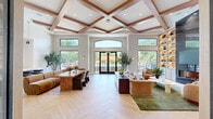 Located at 2107 Stillbrook Ln in Princeton, NJ, this generously sized single-floor amenity space offers a refined blend of modern design and functional comfort. The entrance hallway sets an elegant tone with herringbone light wood flooring, a striking white marble accent wall with a mounted display, and clean architectural lines that guide visitors throughout the space. The expansive living room is a true centerpiece, boasting soaring coffered ceilings with warm wood beam detailing, floor-to-ceiling built-in shelving with integrated lighting, and a dramatic dark marble fireplace surround. Herringbone flooring flows throughout, complemented by large windows that flood the room with natural light and offer views of the surrounding greenery. A pool table and lounge seating areas make this space ideal for social gatherings. The kitchen features warm wood cabinetry, white stone countertops, and a built-in beverage cooler, bathed in natural light from surrounding windows. The dedicated exercise room is well-appointed with a full suite of cardio and strength equipment, vibrant geometric wall murals in bold tones, ceiling fans, and large windows that bring in ample daylight, creating an energizing workout environment.