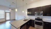 This well-appointed 1-bedroom, 1-bathroom residence in Houston, TX offers a thoughtfully designed single-story layout with modern finishes throughout. The kitchen is a standout feature, showcasing rich dark cabinetry, granite countertops, a subway tile backsplash, and a full suite of stainless steel appliances including a refrigerator with ice maker, electric range, over-the-range microwave, and dishwasher. A peninsula with an undermount sink and pendant lighting above creates a natural transition into the open living area, which features warm wood-style flooring and a ceiling fan. The bedroom is a comfortable retreat with plush carpeting, a ceiling fan, and a large window with blinds that invite natural light. The bathroom is well-equipped with a soaking tub and shower combo featuring tile surround, a dark-cabinet double vanity with granite countertops, wood-style flooring, and direct access to a spacious walk-in closet with built-in shelving. The unit's cohesive design language — dark cabinetry, granite surfaces, and wood-style floors — creates a modern and inviting ambiance throughout.