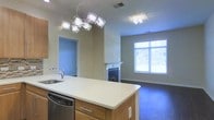 This single-floor space is designed with an open-concept layout that seamlessly connects the kitchen and living areas. The kitchen centers around a large island with a white countertop, warm wood cabinetry, and an integrated stainless steel dishwasher. A striking chandelier with crystal-like pendant fixtures serves as a focal point above the island, adding a contemporary decorative element. The sink is positioned along the perimeter, complemented by neutral-toned walls and standard cabinetry throughout. The adjoining living area features dark flooring that contrasts with the lighter kitchen surfaces, creating a defined yet open transition between spaces. A fireplace with a clean white mantel anchors the living area, providing both a functional and aesthetic centerpiece. A large multi-pane window allows generous natural light to fill the room, offering views of surrounding greenery. A secondary chandelier echoes the kitchen's lighting style, maintaining visual consistency throughout the open floor plan. An additional doorway leads to a separate room, adding to the overall functionality of the layout. The space presents a balance of modern fixtures and practical design elements.